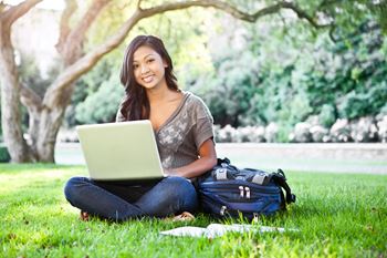 Young female adult working on a laptop while sitting in the grass under a oak tree